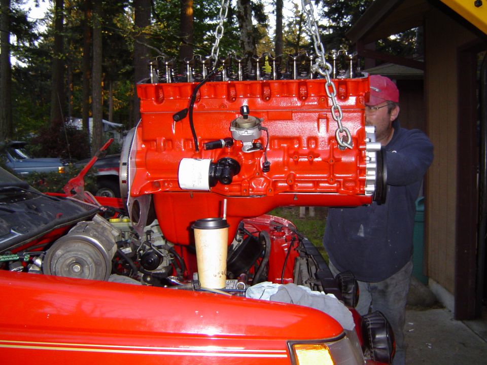 Man in red cap lowering a red-painted inline-six engine by chain hoist into a vehicle