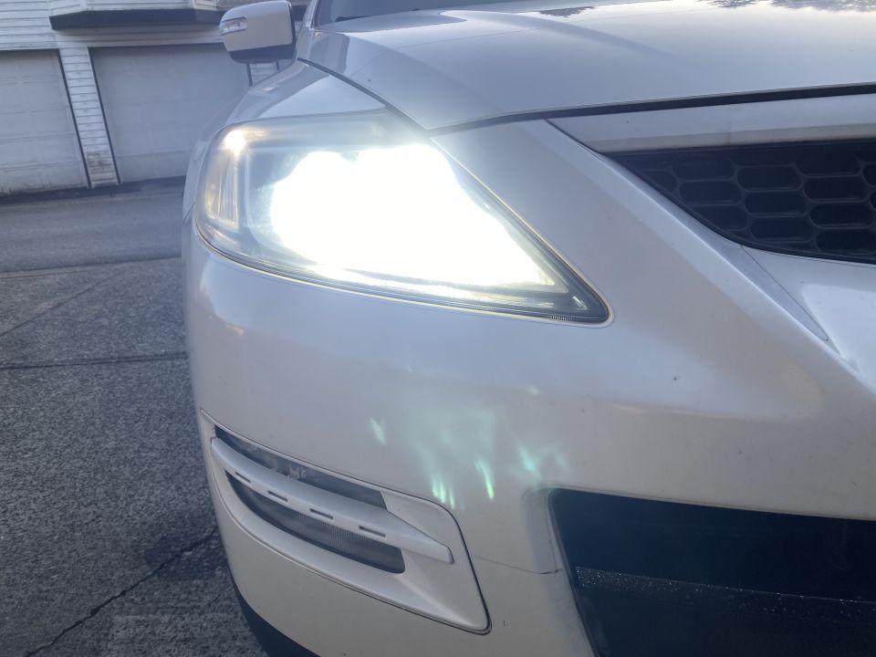 Front passenger side of a white SUV with headlight on, parked on pavement.