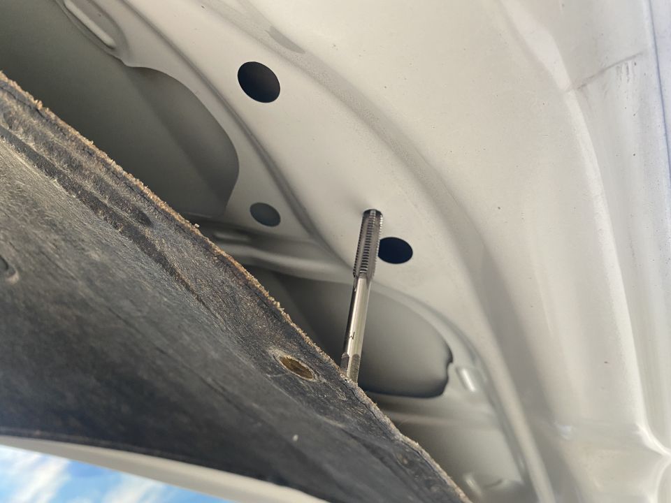 Metal tap tool inserted into a hole on the underside of a white car hood.