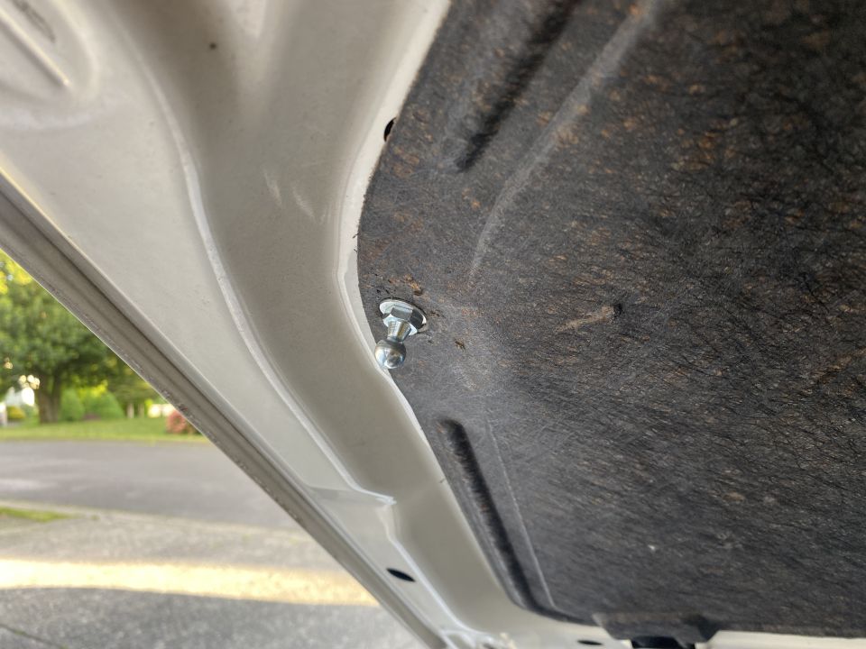 Close-up of bolt holding sound dampening material to underside of white car hood.