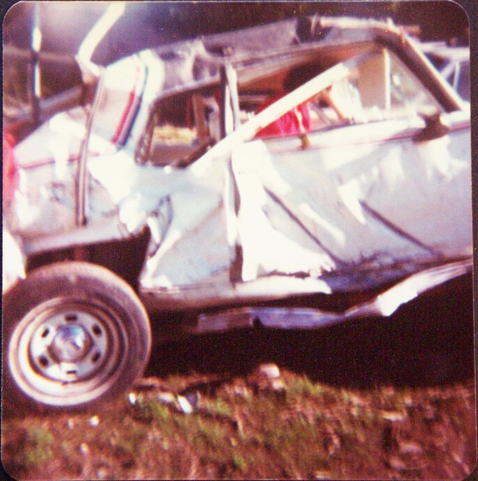 Side view of a heavily crashed white car with crushed body and collapsed roof on grass