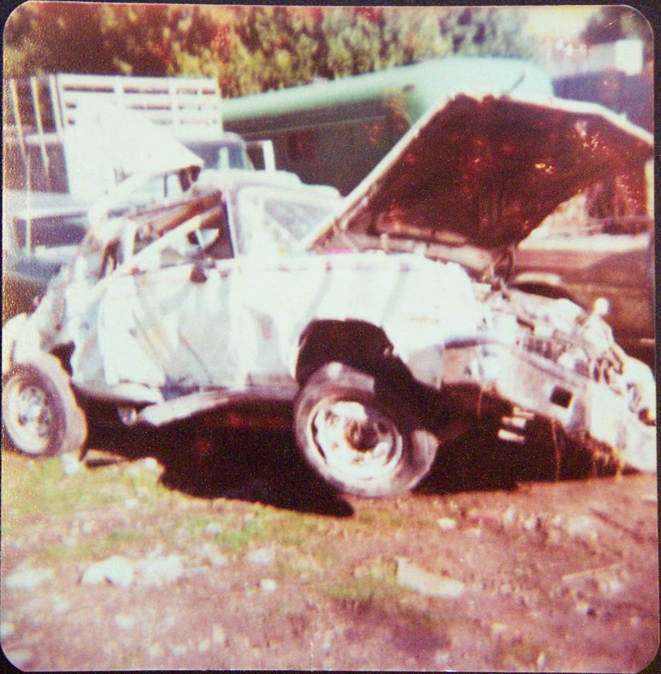 Heavily wrecked light-colored car with crushed body and open hood on dirt lot