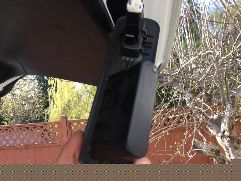 Hand holding black license plate light housing on white vehicle, with fence and trees in background.