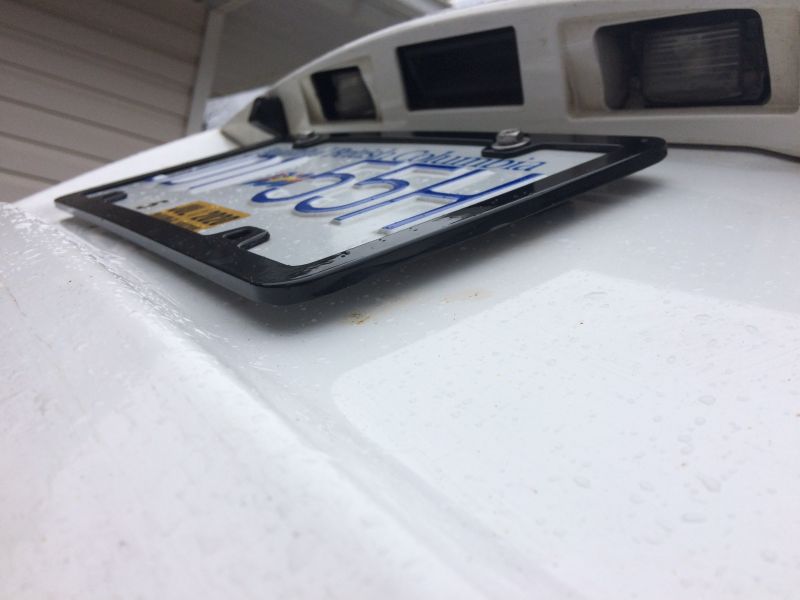License plate frame on a white vehicle with blue lettering and a yellow sticker.