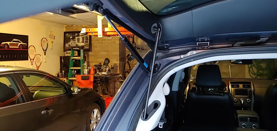 Dark blue SUV rear hatch open, showing interior and gas strut. Garage setting with another car and posters.