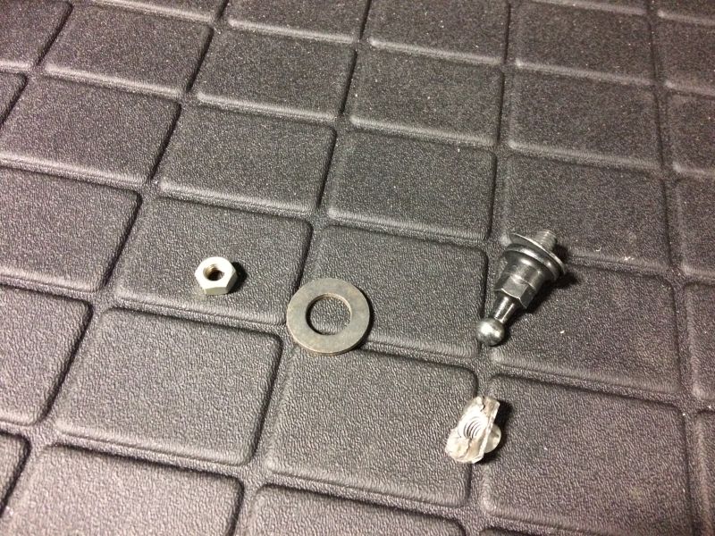 Small metal hardware including a nut, washer, ball stud, and broken metal piece on a textured surface.
