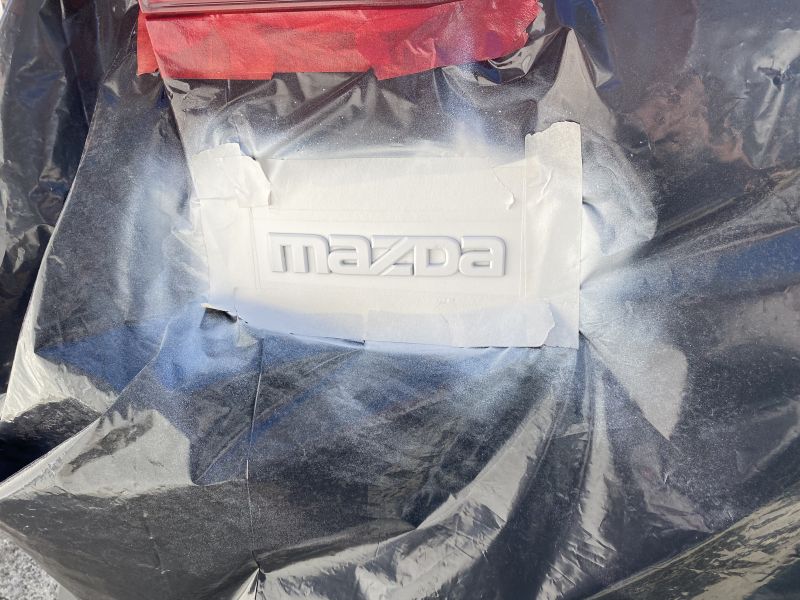White Mazda logo on a car part, covered in plastic sheeting with tape and spray paint overspray.