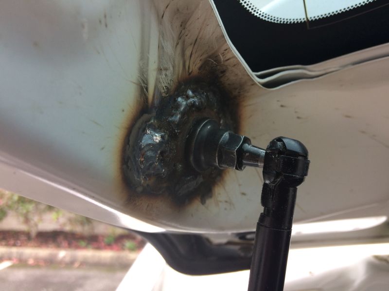 Close-up of a gas strut attached to a welded repair on a white vehicle body panel.