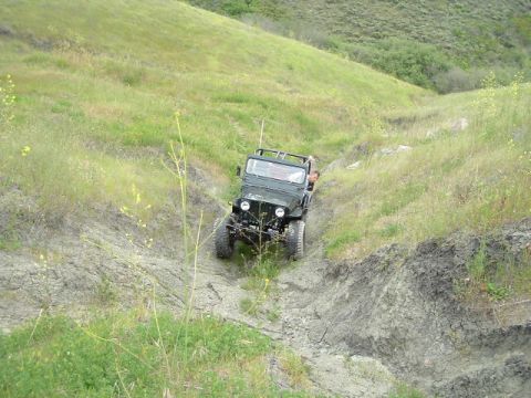 1989 Ford Bronco - A green Jeep CJ-5 navigates a steep, muddy incline surrounded by green, rolling hills.