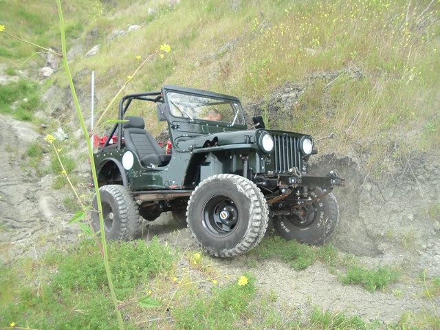 Dark green open-top vintage 4x4 Jeep parked on rocky grassy slope, 3q-front view