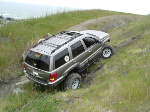 1989 Ford Bronco - A silver Jeep Grand Cherokee navigates a challenging off-road trail with steep inclines and grassy terrain.