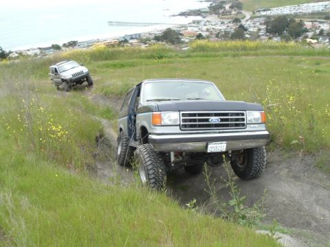 1989 Ford Bronco - Two off-road vehicles navigate a grassy hillside trail near the coast.