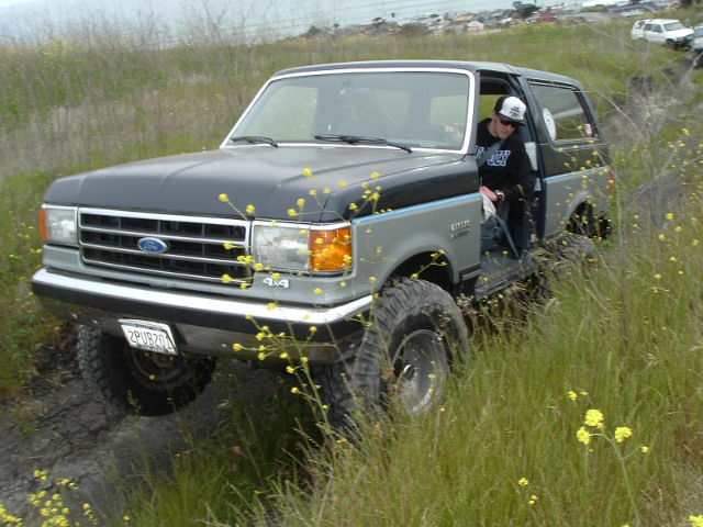 Two-tone gray 1987-91 Ford Bronco 4x4 on mud tires in tall grass and wildflowers, front three-quarter view