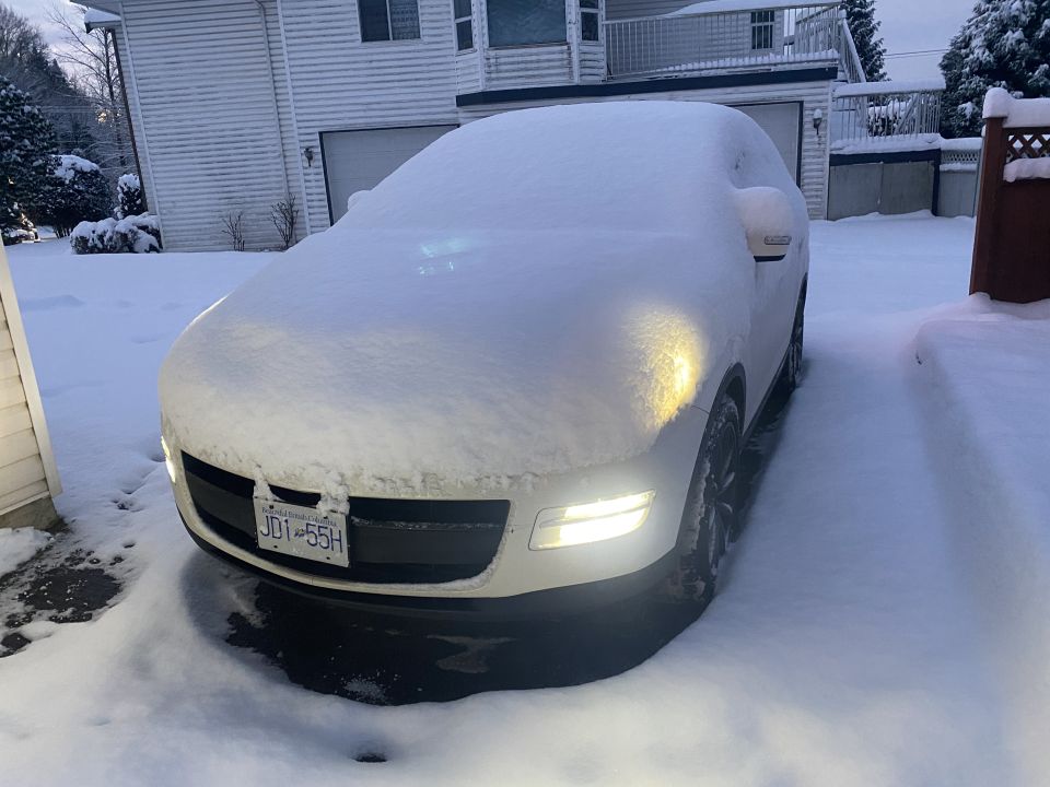 White SUV covered in snow with headlights on in a snowy driveway.