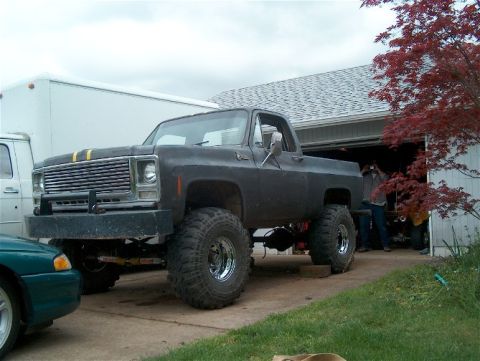 1979 Ford Bronco - A lifted pickup truck with a custom grille and hood stripes sits in a driveway next to a garage.
