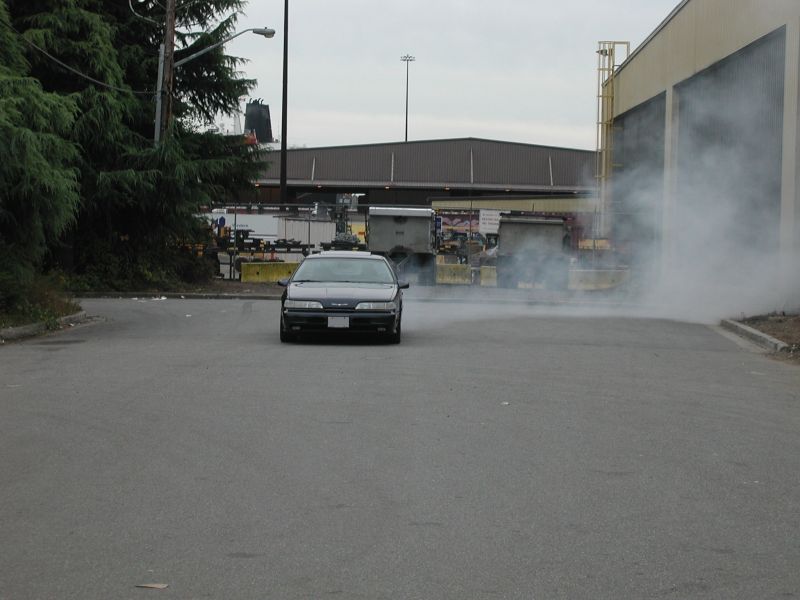 Dark sedan driving and emitting smoke in an industrial area.