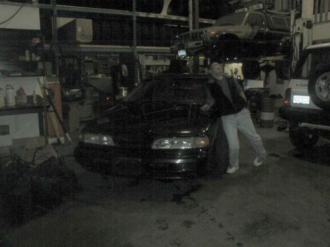 1990 Ford Thunderbird - A man leans on the hood of a black sedan in a dimly lit garage. Another vehicle is lifted on a hydraulic lift in the background.