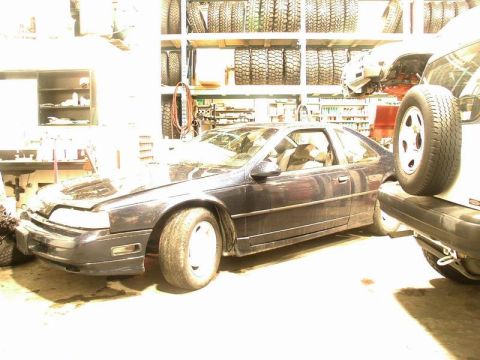 1990 Ford Thunderbird - A dark blue coupe sits in a garage with shelves of tires in the background. The front of the car is visible, showing its headlights and bum…