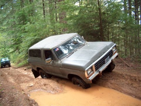 1979 Ford Bronco - A gray 1978 Ford Bronco navigates a muddy trail in a forest, with another vehicle visible in the background.