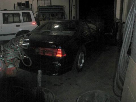 1990 Ford Thunderbird - The rear passenger side of a black coupe is visible in a dimly lit garage. A white SUV is parked to the left of the black car, and a coiled…