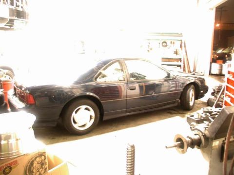1990 Ford Thunderbird - The side profile of a dark blue Ford Thunderbird is visible in a garage setting, parked next to various automotive equipment and supplies.