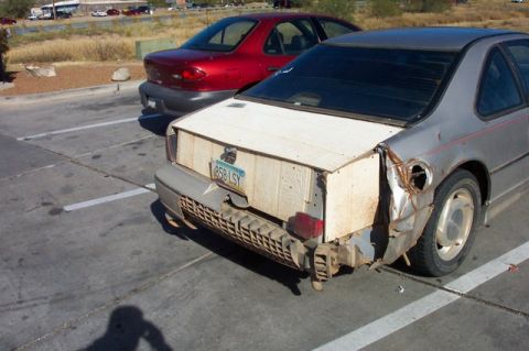 1990 Ford Thunderbird - The rear quarter panel and trunk area of a silver coupe are heavily damaged and crudely repaired with plywood. A red sedan is parked behind…