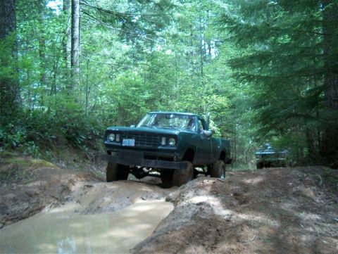 1979 Ford Bronco - A lifted green pickup truck navigates a muddy off-road path surrounded by trees, with another vehicle following.
