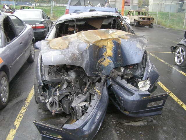 Heavily damaged dark blue sedan with crumpled hood and exposed engine, showing signs of fire damage.