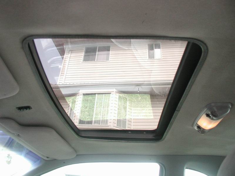 Interior view of a car's sunroof open, showing a house outside. Sun visor and dome light visible.