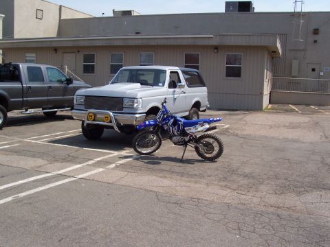 1990 Ford Bronco - A silver Ford Bronco and a gray Ford F-Series truck are parked in a lot with a dirt bike in front of the Bronco.