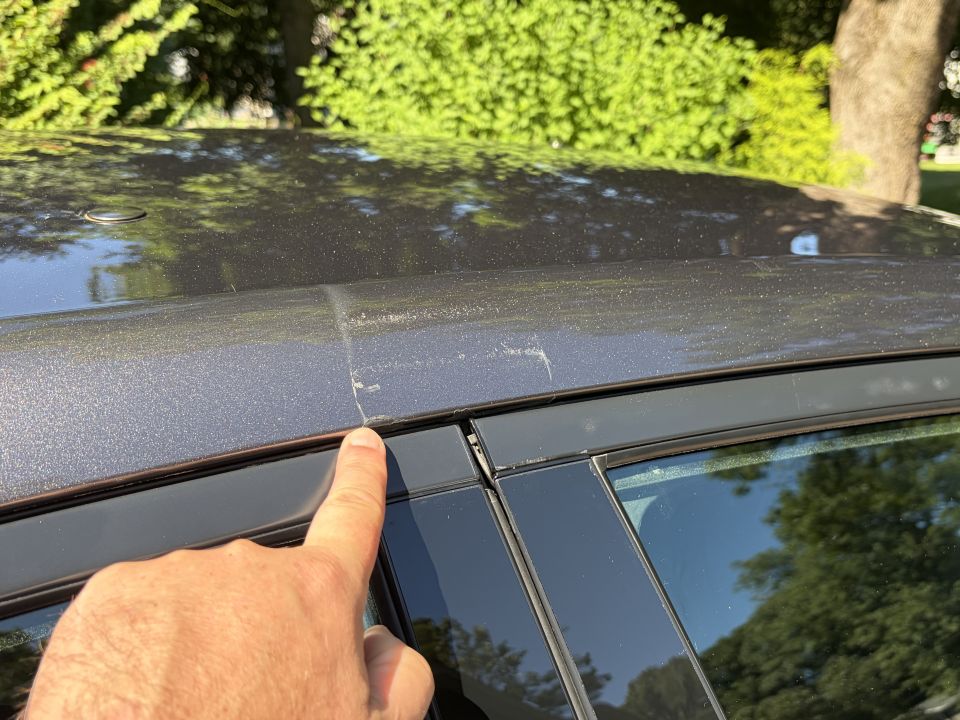 Hand pointing at scuffed paint on the roof rail above a sedan's door glass