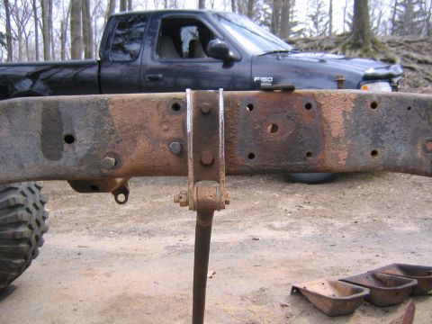 1978 Ford Bronco - A rusty bare truck frame rail held up on a jack stand outdoors, shown side-on with a fresh weld along a bracket and old body-mount holes an…