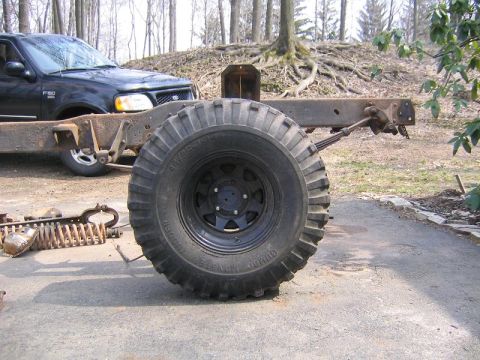 1978 Ford Bronco - A large Jumbo Monster Mudder off-road tire mounted on a black eight-spoke steel wheel standing upright on a driveway, with a bare rusty tru…