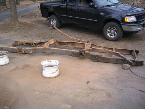 1978 Ford Bronco - A completely stripped, rust-covered truck frame lying bare on a concrete driveway with its rear leaf springs still attached, propped up on…