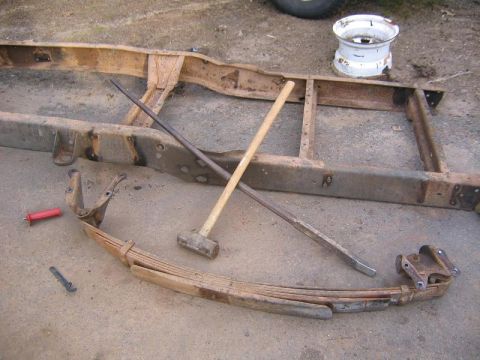 1978 Ford Bronco - A rusty multi-leaf spring pack with its shackle and hanger brackets removed and laid on a concrete driveway, next to a sledgehammer and a l…