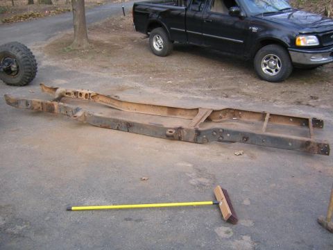 1978 Ford Bronco - A bare, rusty truck ladder frame lying flat on a gravel driveway, stripped of body, suspension and drivetrain so the boxed rails, crossmemb…