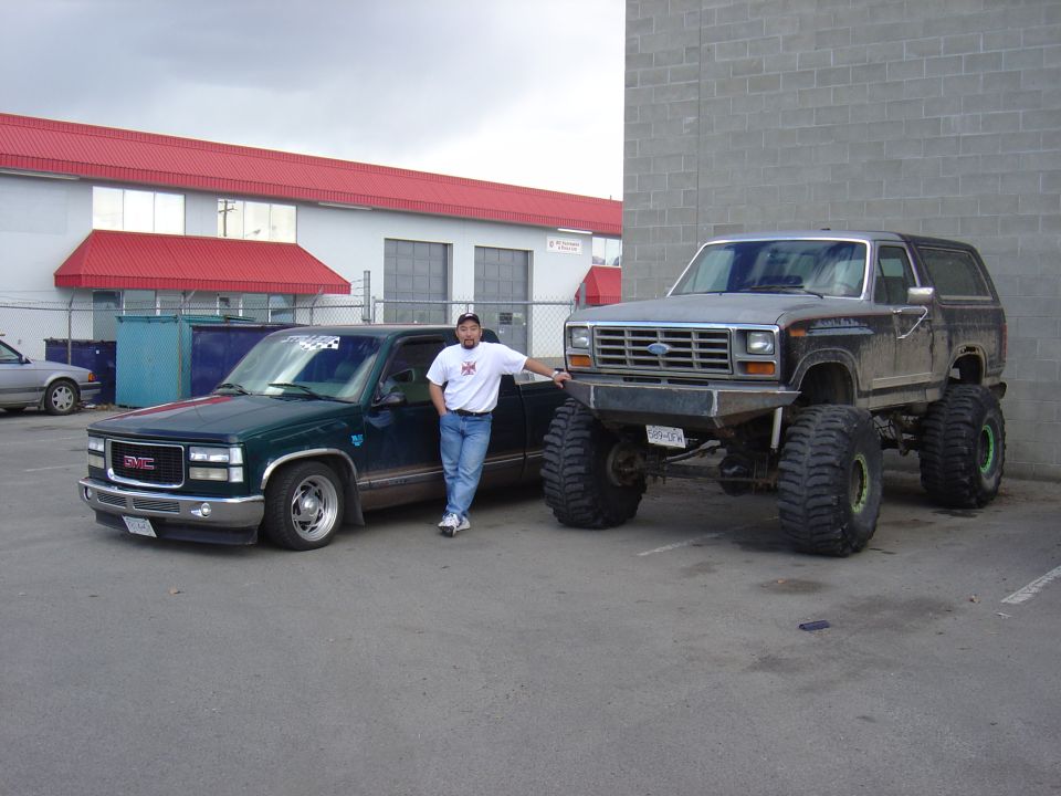Lifted brown Ford Bronco on big mud tires parked beside a lowered green GMC pickup, man standing between them