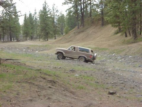 1989 Ford Bronco - A brown Ford Bronco navigates a rough, off-road terrain with trees and a hill in the background.
