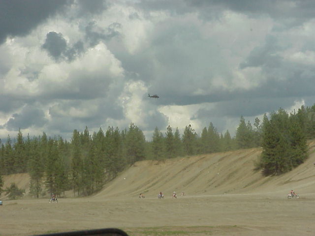 Dirt bikes ride on a sandy hill under a cloudy sky with a helicopter flying