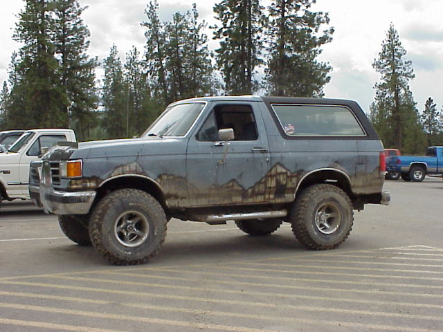 1989 Ford Bronco parked next to a white pickup truck, with a blue pickup truck in the background