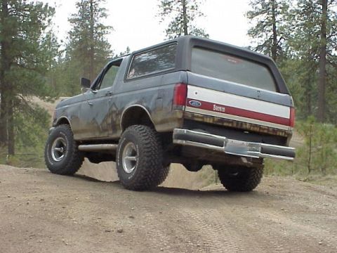 1989 Ford Bronco - This lifted Ford Bronco is seen from the rear on a dirt road, with off-road tires and a distinctive red and white stripe on its tailgate.
