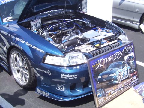 2002 Ford Mustang - A modified blue Ford Mustang is displayed with its engine exposed, alongside a promotional poster.