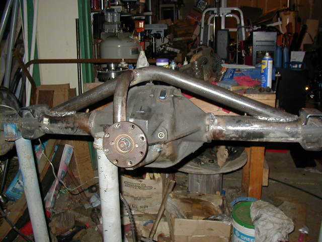 Bare rear axle housing with welded tubular links on a jack stand in a cluttered shop