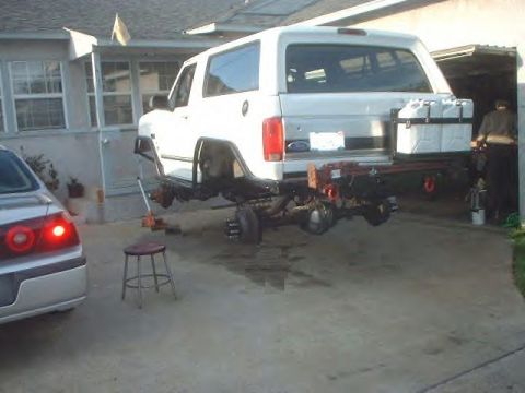 1979 Ford Bronco - White SUV chassis exposed as it is lifted for maintenance, with spare fuel containers mounted at the rear.