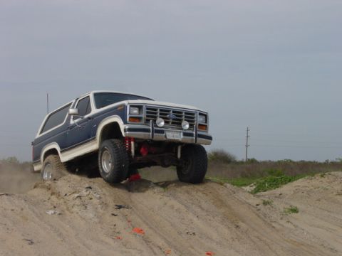 1986 Ford Bronco - This lifted Ford Bronco is shown off-roading on a sandy incline under a cloudy sky.