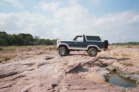 1986 Ford Bronco - A blue and white 1986 Ford Bronco is parked on a rocky riverbed under a partly cloudy sky.