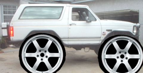 1990 Ford Bronco - Side profile of a white square-body 1980s Ford Bronco under a carport, with an oversized pair of silver multi-spoke aftermarket wheels digi…