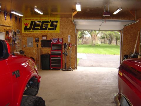 1978 Ford Bronco - Interior view of a garage containing two red vehicles and various tools, with the garage door open to a yard.