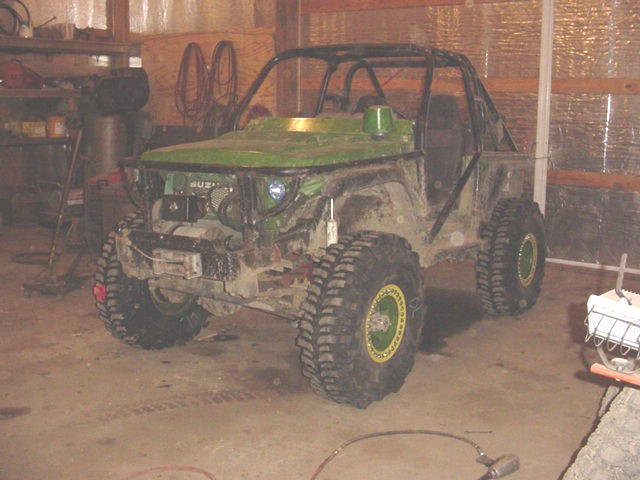 Green off-road vehicle with roll cage, large tires, and exposed engine in a garage