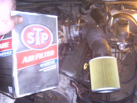 1991 Ford LTD Crown Victoria - A hand holds an STP air filter box, part number SA6543, over an open engine bay while a new round yellow-pleated air filter element sits on…